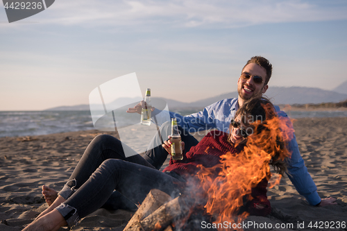 Image of Young Couple Sitting On The Beach beside Campfire drinking beer