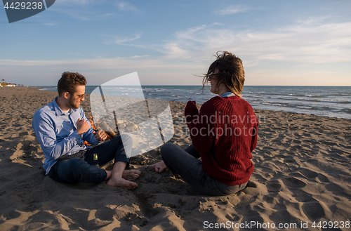 Image of Young Couple Sitting On The Beach beside Campfire drinking beer