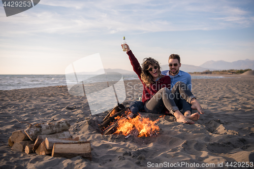 Image of Young Couple Sitting On The Beach beside Campfire drinking beer