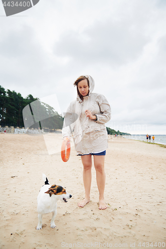 Image of Young woman in jacket playing with dog on beach