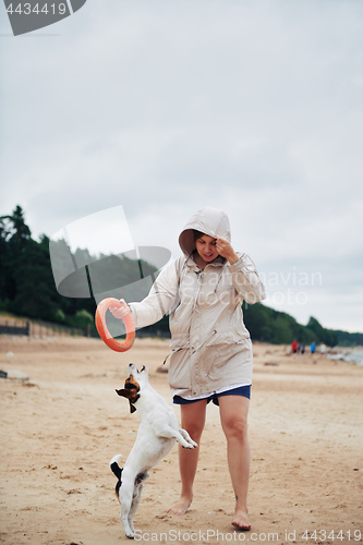 Image of Young woman in jacket playing with dog on beach