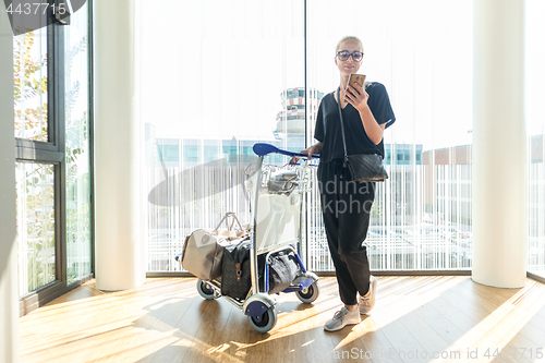 Image of Casual woman on airport terminal talking on the cell phone, waiting for flight departure with luggage on trolley cart.