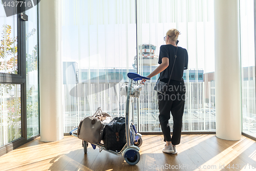 Image of Casual woman on airport terminal talking on the cell phone, waiting for flight departure with luggage on trolley cart.