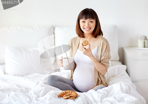 Image of happy pregnant woman eating cookie in bed at home