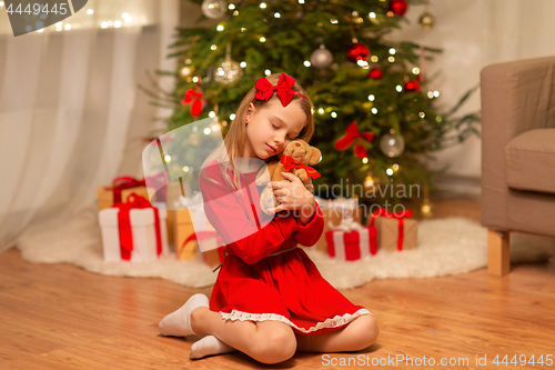 Image of girl in red dress hugging teddy bear at home