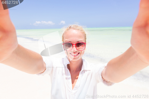 Image of Young woman wearing white beach tunic taking selfie on tropical white sandy beach.