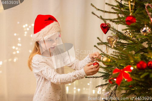 Image of happy girl in santa hat decorating christmas tree
