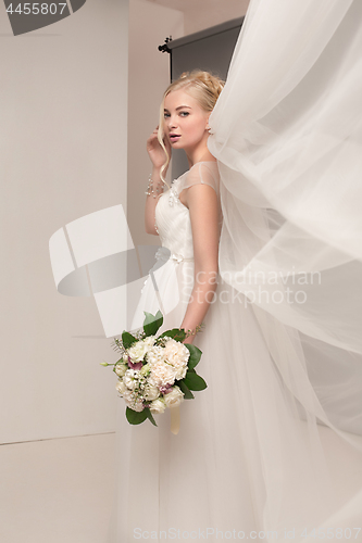 Image of Bride in beautiful dress standing indoors in white studio interior like at home. Trendy wedding style shot. Young attractive caucasian model like a bride tender looking.
