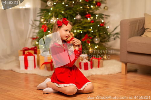 Image of girl in red dress hugging teddy bear at home