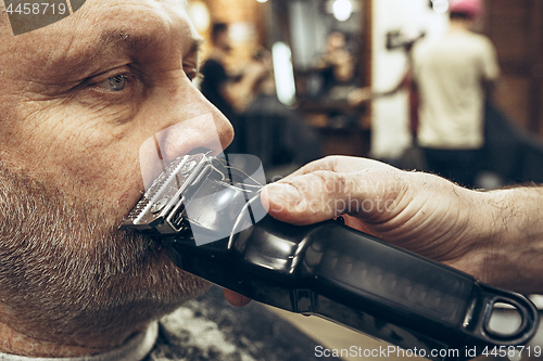 Image of Close-up side view portrait of handsome senior bearded caucasian man getting beard grooming in modern barbershop.