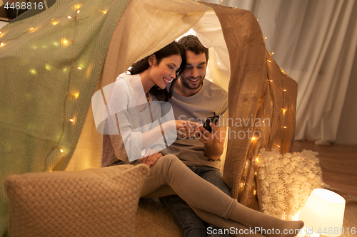 Image of happy couple with smartphone in kids tent at home