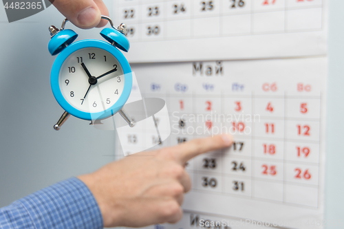 Image of A hand holds a small alarm clock, in the background a hand points to long weekends and holidays on a wall calendar