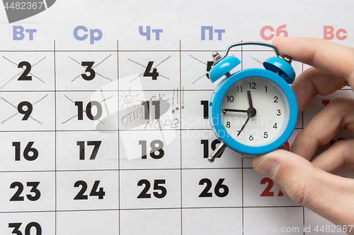 Image of A hand holds a small alarm clock, in the background is a calendar with the day marked and the word vacation