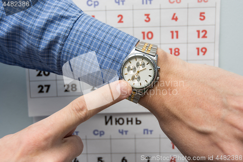 Image of Office specialist\'s hand points to a wristwatch, in the background is a wall calendar