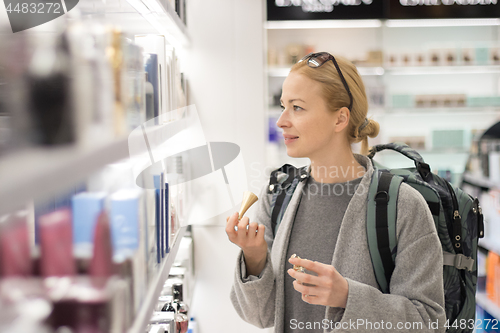 Image of Blond young female traveler wearing travel backpack choosing perfume in airport duty free store.