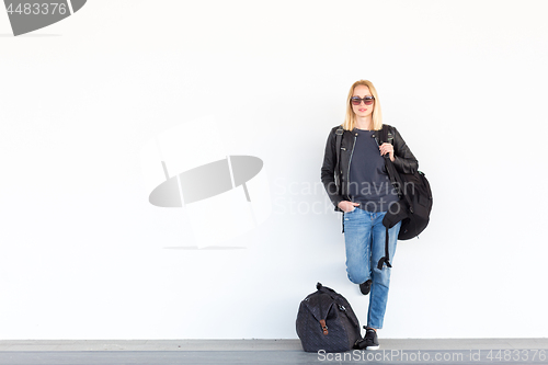 Image of Fashionable young woman standing and waiting against plain white wall on the station whit travel bag by her side.