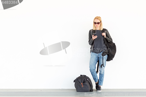 Image of Fashionable young woman using her mobile phone while standing and waiting against plain white wall on the station whit travel bag by her side.