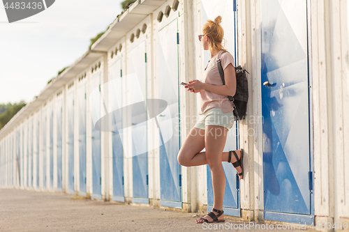 Image of Blonde young female traveler wearing summer style clothing, holding mobile phone, against retro blue beach dressing rooms at summer time vacation.