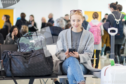 Image of Cheerful female traveler smiling, looking at camera while reading on her cell phone while waiting to board a plane at departure gates at airport terminal.