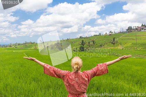 Image of Relaxed fashionable caucasian woman wearing red asian style kimono, arms rised to sky, enjoying pure nature of beautiful green rice fields on Bali.