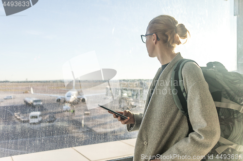 Image of Casually dressed female traveler at airport, holding smart phone device, looking through the airport gate windows at planes on airport runway