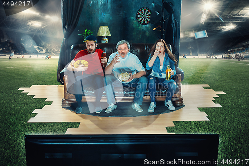 Image of Soccer football fans sitting on the sofa and watching TV in the middle of a football field.