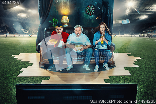 Image of Soccer football fans sitting on the sofa and watching TV in the middle of a football field.