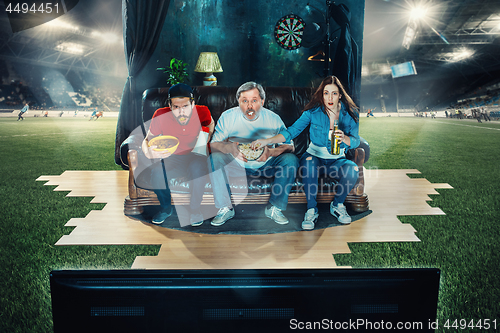 Image of Soccer football fans sitting on the sofa and watching TV in the middle of a football field.