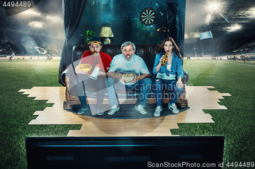 Image of Soccer football fans sitting on the sofa and watching TV in the middle of a football field.