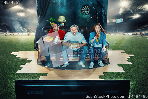 Image of Soccer football fans sitting on the sofa and watching TV in the middle of a football field.