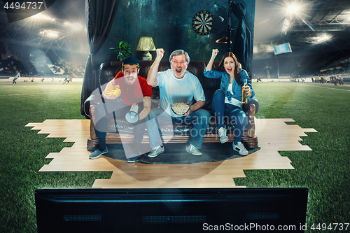 Image of Soccer football fans sitting on the sofa and watching TV in the middle of a football field.