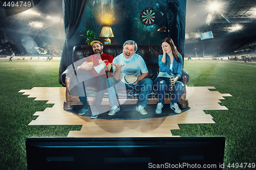 Image of Soccer football fans sitting on the sofa and watching TV in the middle of a football field.