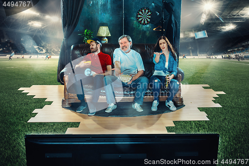 Image of Soccer football fans sitting on the sofa and watching TV in the middle of a football field.