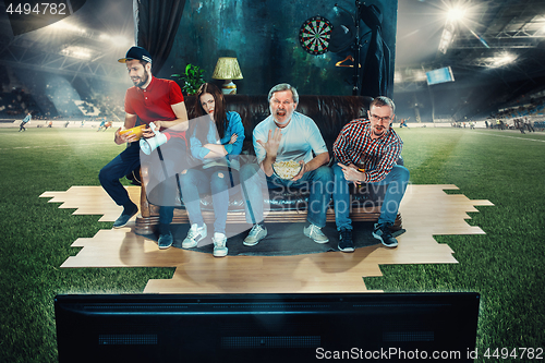 Image of Soccer football fans sitting on the sofa and watching TV in the middle of a football field.