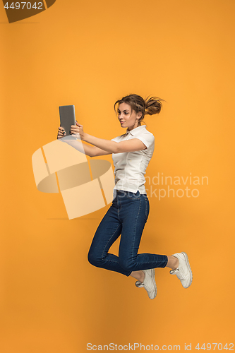 Image of Image of young woman over orange background using laptop computer or tablet gadget while jumping.