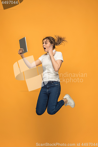 Image of Image of young woman over orange background using laptop computer or tablet gadget while jumping.