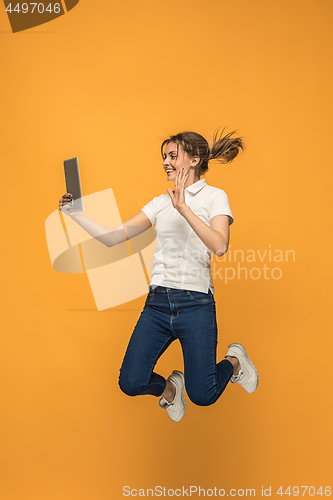 Image of Image of young woman over orange background using laptop computer or tablet gadget while jumping.