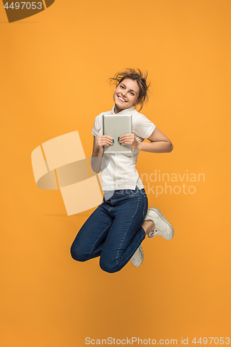 Image of Image of young woman over orange background using laptop computer or tablet gadget while jumping.