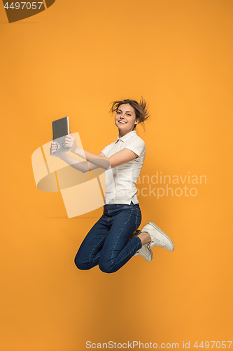 Image of Image of young woman over orange background using laptop computer or tablet gadget while jumping.