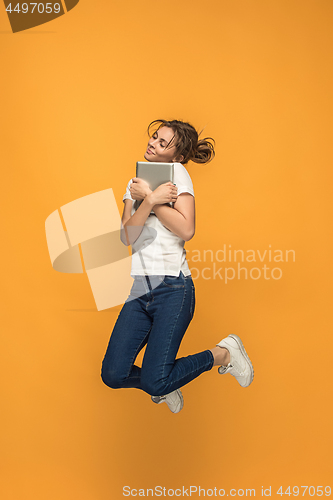 Image of Image of young woman over orange background using laptop computer or tablet gadget while jumping.