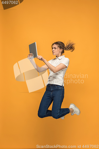 Image of Image of young woman over orange background using laptop computer or tablet gadget while jumping.
