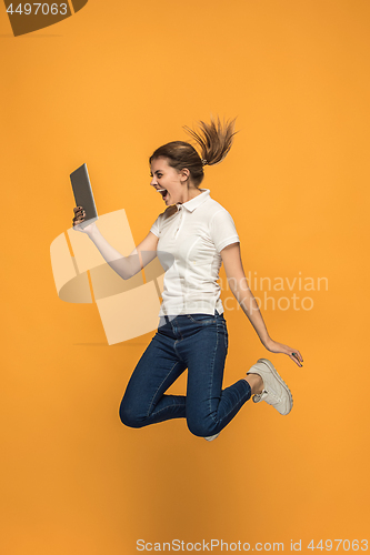 Image of Image of young woman over orange background using laptop computer or tablet gadget while jumping.