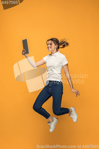 Image of Image of young woman over orange background using laptop computer or tablet gadget while jumping.