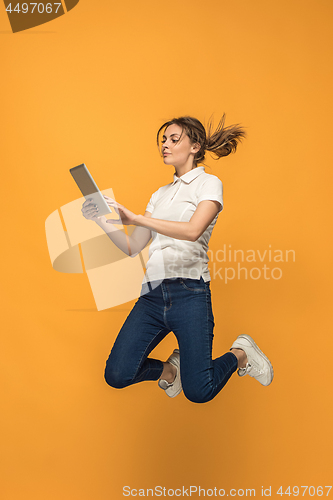 Image of Image of young woman over orange background using laptop computer or tablet gadget while jumping.