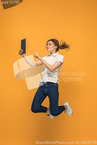 Image of Image of young woman over orange background using laptop computer or tablet gadget while jumping.