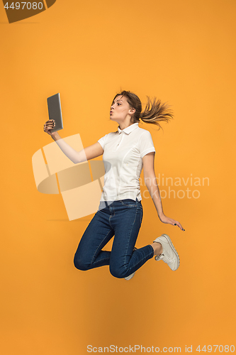 Image of Image of young woman over orange background using laptop computer or tablet gadget while jumping.