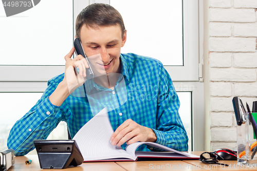 Image of Joyful specialist in the office is talking on the phone and leafing through documents