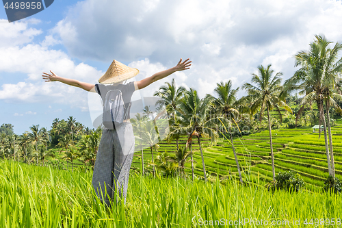 Image of Relaxed fashionable female traveler wearing small backpack and traditional asian paddy hat, arms rised to sky, enjoying pure nature at beautiful green rice fields and terraces on Bali island