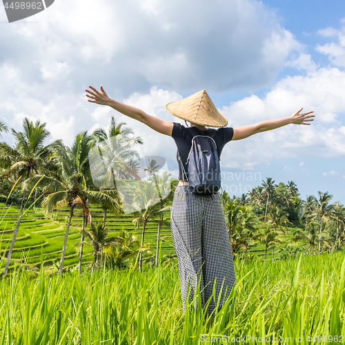 Image of Relaxed fashionable female traveler wearing small backpack and traditional asian paddy hat, arms rised to sky, enjoying pure nature at beautiful green rice fields and terraces on Bali island