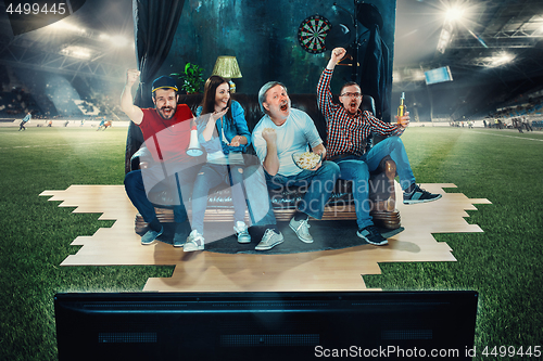 Image of Soccer football fans sitting on the sofa and watching TV in the middle of a football field.